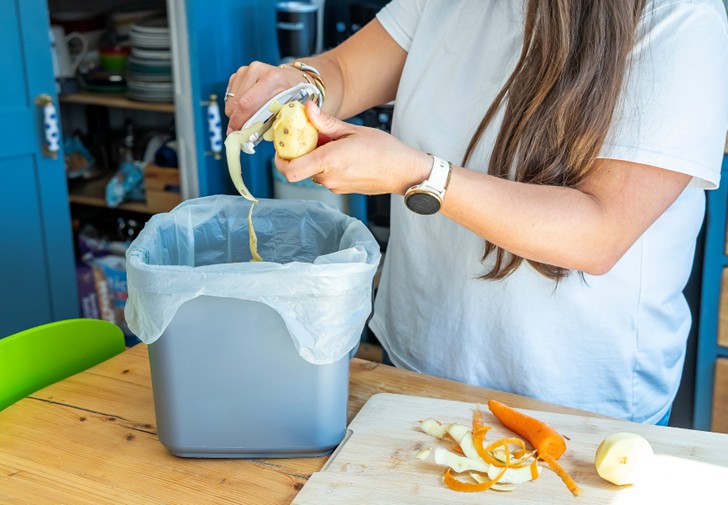 A woman standing in a kitchen peeling fruit into a food waste caddy.